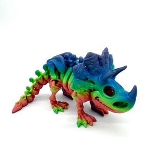 May include: A rainbow-colored, 3D printed dinosaur skeleton toy. The dinosaur is a triceratops with a large frill and three horns. The toy is made of plastic and is about 6 inches long.