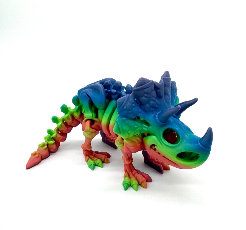May include: A rainbow-colored, 3D printed dinosaur skeleton toy. The dinosaur is a triceratops with a large frill and three horns. The toy is made of plastic and is about 6 inches long.