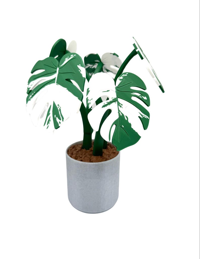 K&ouml;nnte beinhalten: Eine k&uuml;nstliche Monstera-Pflanze in einem Topf mit gro&szlig;en, gr&uuml;n-wei&szlig; panaschierten Bl&auml;ttern. Die Pflanze steht in einem hellgrauen, zylindrischen Topf, der mit braunem Material gef&uuml;llt ist, um Erde zu simulieren. Die Bl&auml;tter haben ein charakteristisches Lochmuster.