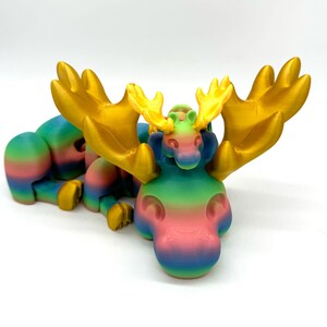 Op de afbeelding: Een regenboogkleurig 3D-geprint elandbeeldje met grote gouden geweien. Het eland ligt met zijn hoofd opzij gedraaid.