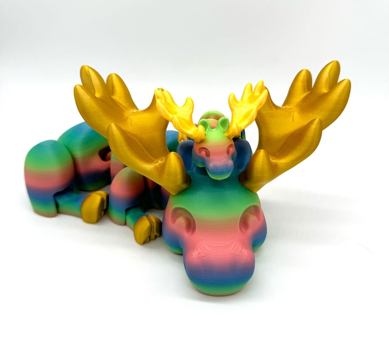 Op de afbeelding: Een regenboogkleurig 3D-geprint elandbeeldje met grote gouden geweien. Het eland ligt met zijn hoofd opzij gedraaid.