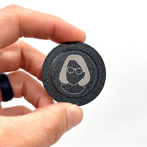 May include: A black round button with a silver outline and a silver silhouette of a person wearing glasses.