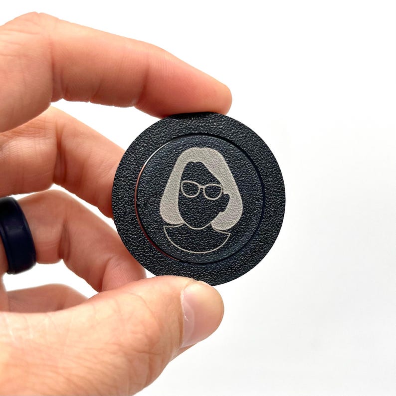 May include: A black round button with a silver outline and a silver silhouette of a person wearing glasses.