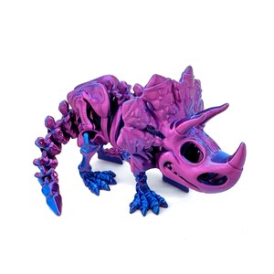 May include: A purple and blue 3D printed dinosaur skeleton figurine. The dinosaur is a Triceratops with a large frill and three horns. The figurine is detailed and has a textured finish.