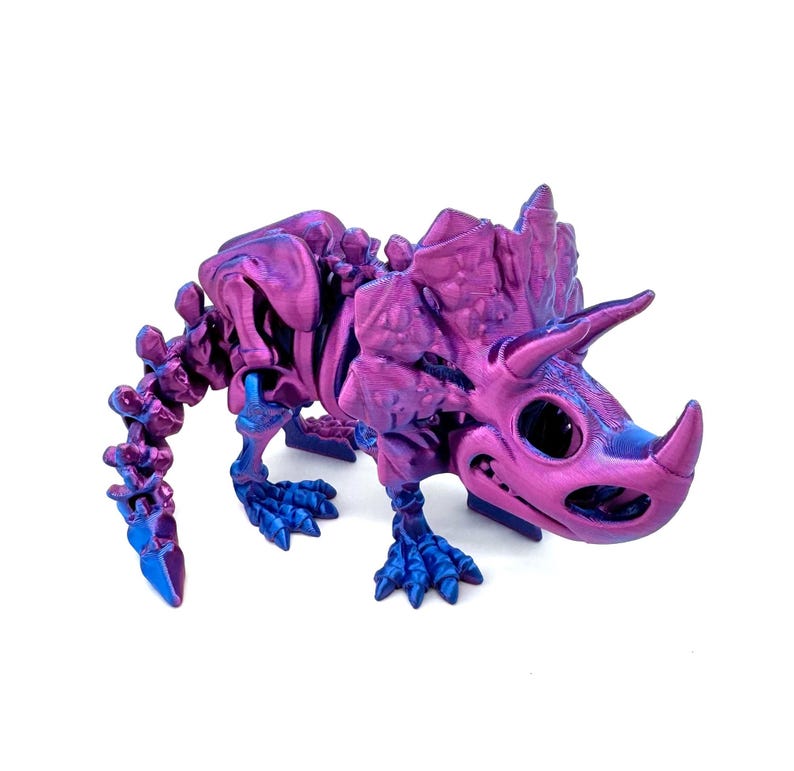 May include: A purple and blue 3D printed dinosaur skeleton figurine. The dinosaur is a Triceratops with a large frill and three horns. The figurine is detailed and has a textured finish.