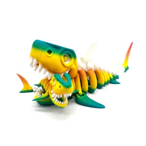 May include: A 3D printed skeleton of a shark with a smaller shark skeleton inside its mouth. The larger shark is painted in shades of green, yellow, and orange. The smaller shark is painted in shades of green and yellow.