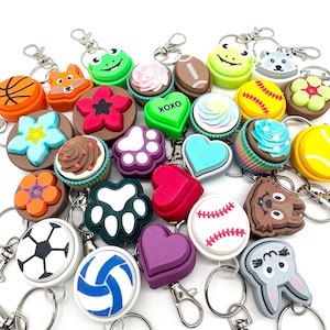 Puede incluir: Una colección de llaveros coloridos con varios diseños, incluyendo animales, deportes, corazones y huellas de patas. Los llaveros están hechos de plástico y tienen un efecto 3D.