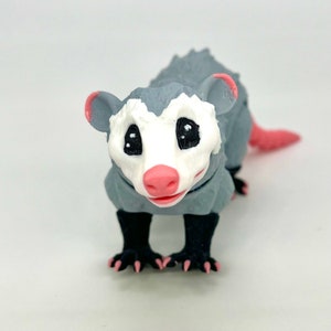 May include: A gray and white 3D printed opossum figurine with black eyes and a pink nose. The opossum is standing on all fours with its tail curled up behind it.