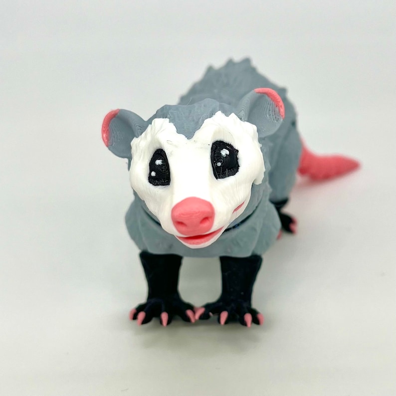 May include: A gray and white 3D printed opossum figurine with black eyes and a pink nose. The opossum is standing on all fours with its tail curled up behind it.