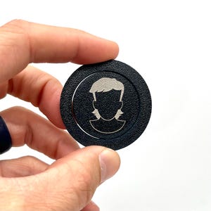 May include: A black round token with a silver silhouette of a person with a mullet hairstyle.