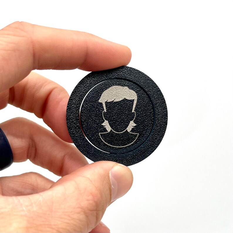 May include: A black round token with a silver silhouette of a person with a mullet hairstyle.