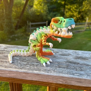 May include: A colorful 3D printed dinosaur skeleton toy. The dinosaur is green, orange, and white. It is posed in a standing position with its mouth open.
