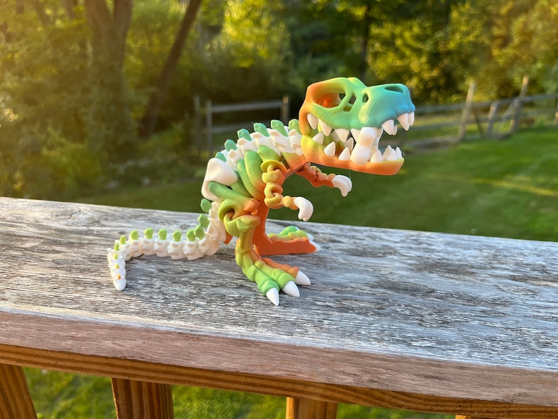 May include: A colorful 3D printed dinosaur skeleton toy. The dinosaur is green, orange, and white. It is posed in a standing position with its mouth open.