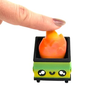 May include: A 3D-printed toy fire pit with a flame. The fire pit is green and yellow with a black border and wheels. The flame is orange and yellow. The fire pit has a smiling face.