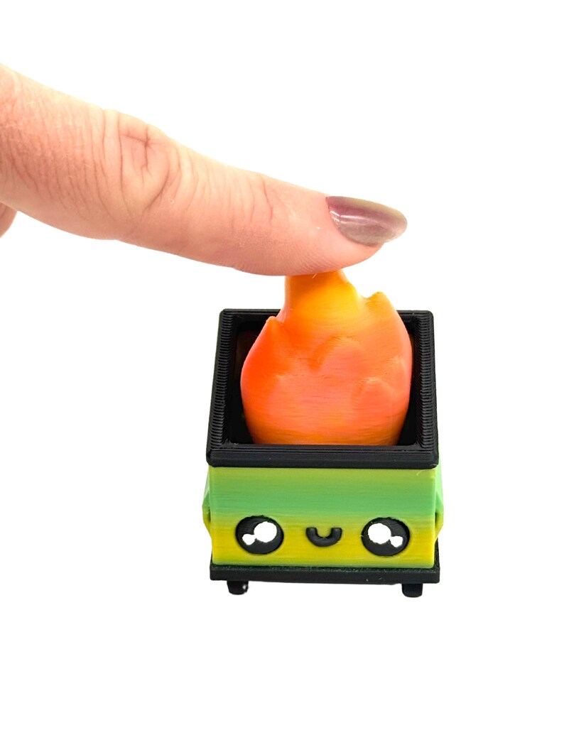 May include: A 3D-printed toy fire pit with a flame. The fire pit is green and yellow with a black border and wheels. The flame is orange and yellow. The fire pit has a smiling face.