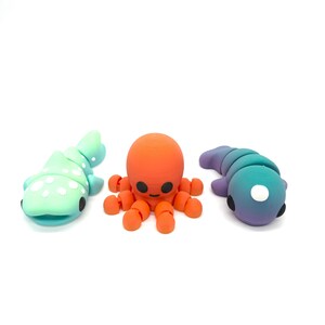 May include: Three colourful clay figurines of sea creatures: a light blue and white whale shark, an orange octopus with eight tentacles, and a purple and blue fish.
