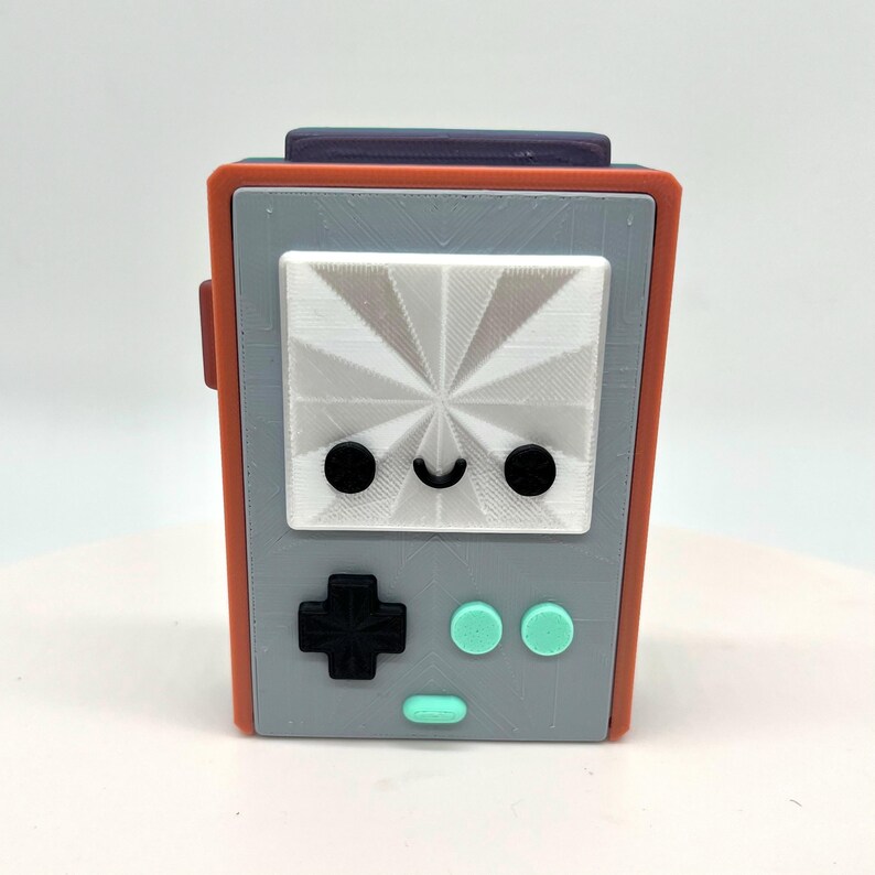 May include: A small, gray and orange handheld game console with a white screen and a smiley face. The console has a black cross-shaped button and two teal round buttons.