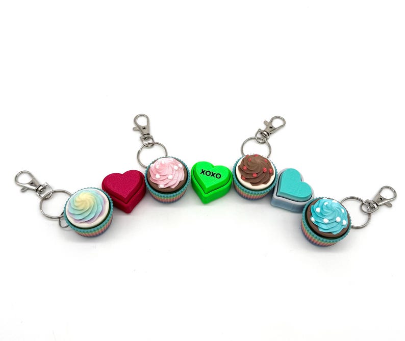 May include: A set of six keychains featuring different cupcake and heart designs. The keychains are made of colorful plastic and have silver metal rings. The cupcake designs include a pink cupcake with white frosting, a blue cupcake with white frosting, a brown cupcake with chocolate frosting, and a rainbow cupcake with white frosting. The heart designs include a red heart and a green heart with the word "XOXO" printed on it.