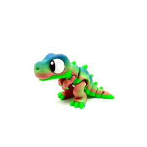 May include: A small, colourful, 3D printed dinosaur toy with a green, blue and pink gradient. The dinosaur has large, white eyes and a playful expression.