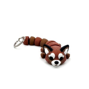 May include: A red panda keyring with a brown and black segmented tail. The red panda has white facial markings and a black nose.