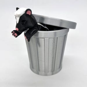 May include: A small black and white skunk figurine is peeking out of a silver trash can with a lid. The skunk has pink feet and a white face.
