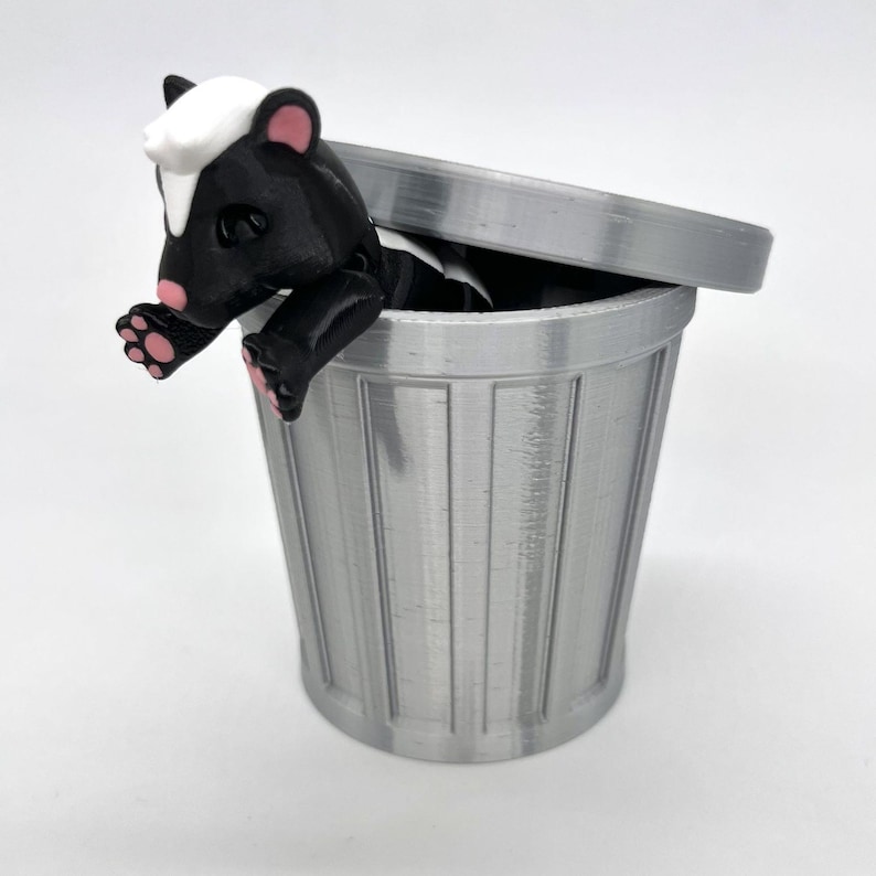 May include: A small black and white skunk figurine is peeking out of a silver trash can with a lid. The skunk has pink feet and a white face.