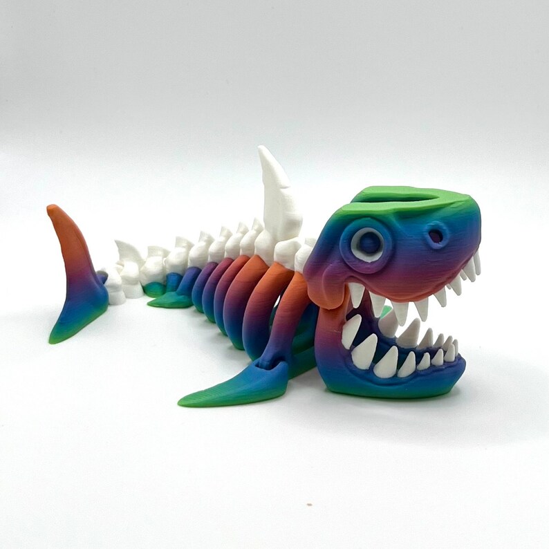 May include: A rainbow-colored 3D printed shark skeleton with a large open mouth and white teeth. The shark has a white dorsal fin and a rainbow-colored tail fin.