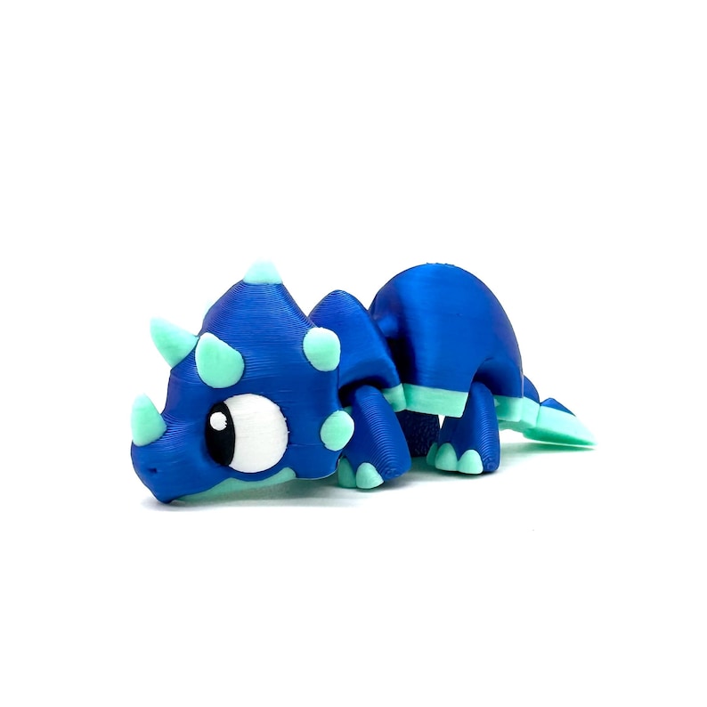 May include: A blue and green 3D printed toy triceratops dinosaur with large eyes. The dinosaur is posed in a walking position with its head down.