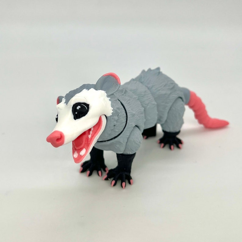 May include: A gray and pink plastic toy opossum with a white face and large black eyes. The opossum has a wide open mouth and pink teeth.