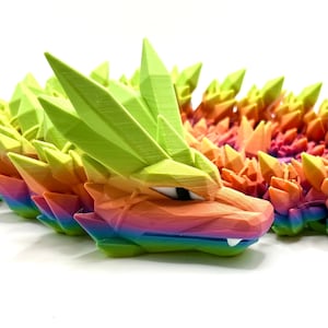 May include: A rainbow-colored, 3D printed dragon figurine with a spiky, geometric design. The dragon's head is facing forward with its mouth slightly open, revealing sharp teeth.