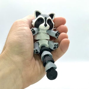 May include: A white and gray articulated raccoon figurine with a black and gray segmented tail. The figurine has black eyes and a black nose.