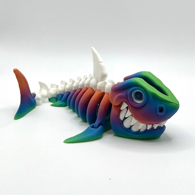 May include: A rainbow colored 3D printed shark skeleton with a toothy grin. The shark is made of plastic and has a white dorsal fin.