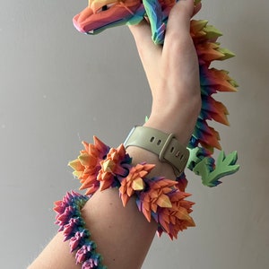 May include: A rainbow-colored 3D printed dragon figurine with a spiky, crystal-like design. The dragon is being held by a hand wearing a matching rainbow-colored bracelet.