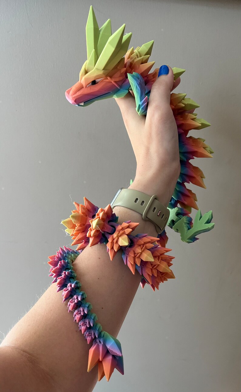 May include: A rainbow-colored 3D printed dragon figurine with a spiky, crystal-like design. The dragon is being held by a hand wearing a matching rainbow-colored bracelet.