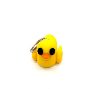 May include: A yellow rubber duck keyring with black eyes and an orange beak. The duck is attached to a silver key ring.