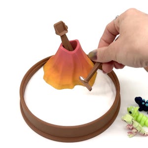 May include: A miniature volcano toy with a brown ring base, a gradient orange and pink volcano, and two brown tools. The volcano is set in a white surface. Small, colorful toy creatures are in the background.