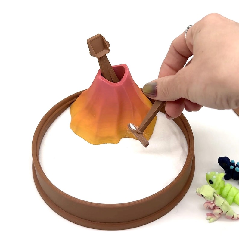 May include: A miniature volcano toy with a brown ring base, a gradient orange and pink volcano, and two brown tools. The volcano is set in a white surface. Small, colorful toy creatures are in the background.