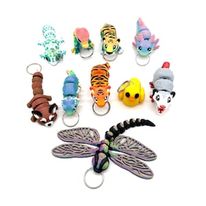 May include: A set of ten colourful animal keychains, including a red panda, a tiger, a dragonfly, a caterpillar, a gecko, an axolotl, a duck, a possum, and two more tigers. The keychains are made of polymer clay and are hand-painted.