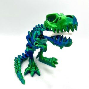 May include: A green and blue 3D printed dinosaur skeleton toy with a white bone in its mouth.