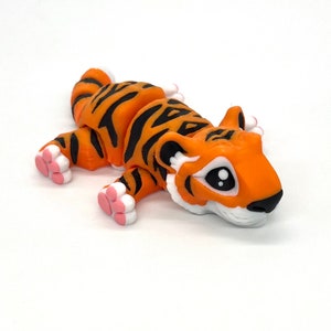 K&ouml;nnte beinhalten: Eine kleine orange-schwarze Tigerfigur mit wei&szlig;en Pfoten und schwarzen Augen. Der Tiger liegt auf dem Bauch mit dem Kopf zur Seite gedreht.