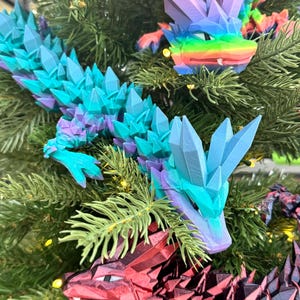 May include: Three colorful, crystal-like dragon ornaments hanging on a Christmas tree. The dragons are red, blue, and purple with iridescent accents.