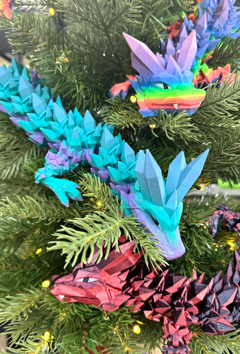 May include: Three colorful, crystal-like dragon ornaments hanging on a Christmas tree. The dragons are red, blue, and purple with iridescent accents.