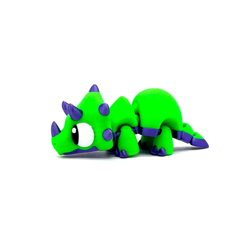 May include: A green and purple toy dinosaur with a large eye and a small horn. The dinosaur is made of plastic and has a cartoonish appearance.
