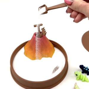 May include: A miniature volcano toy with a brown base and a colorful orange and pink cone. White sand is being poured onto the volcano from a small brown shovel. Small dinosaur figurines are scattered around the base.