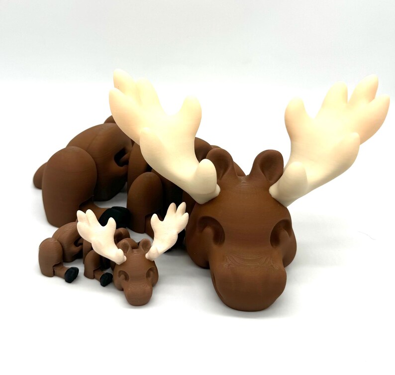Op de afbeelding: Een set van drie bruine eland-beeldjes met witte gewei. De grootste eland ligt met zijn kop op zijn voorpoten. De twee kleinere elanden liggen naast de grootste.