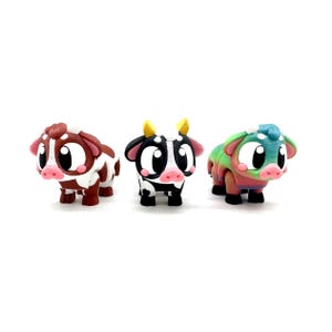 May include: Three small cow figurines, one brown and white, one black and white, and one multi-coloured. Each cow has large eyes and a friendly expression.