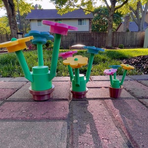 May include: A set of five colorful flower-shaped planters with green stems and brown pots. The planters are arranged in a row on a brick patio.