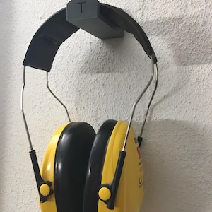 May include: Yellow and black earmuffs hanging on a gray wall mount. The earmuffs have a black headband and silver wire supports. The wall mount is gray and has the letter "T" on it. The wall is white and textured.