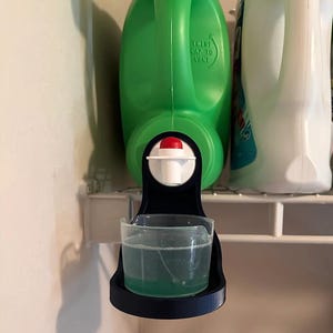 May include: A green laundry detergent bottle with a dispenser attached to a black plastic holder. The dispenser has a clear measuring cup with green liquid.