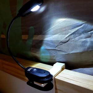 May include: A black LED reading light with a flexible neck, clipped to a wooden surface. The light is on, illuminating the surrounding area. The base of the light has the text "MIGHT BRIGHT".
